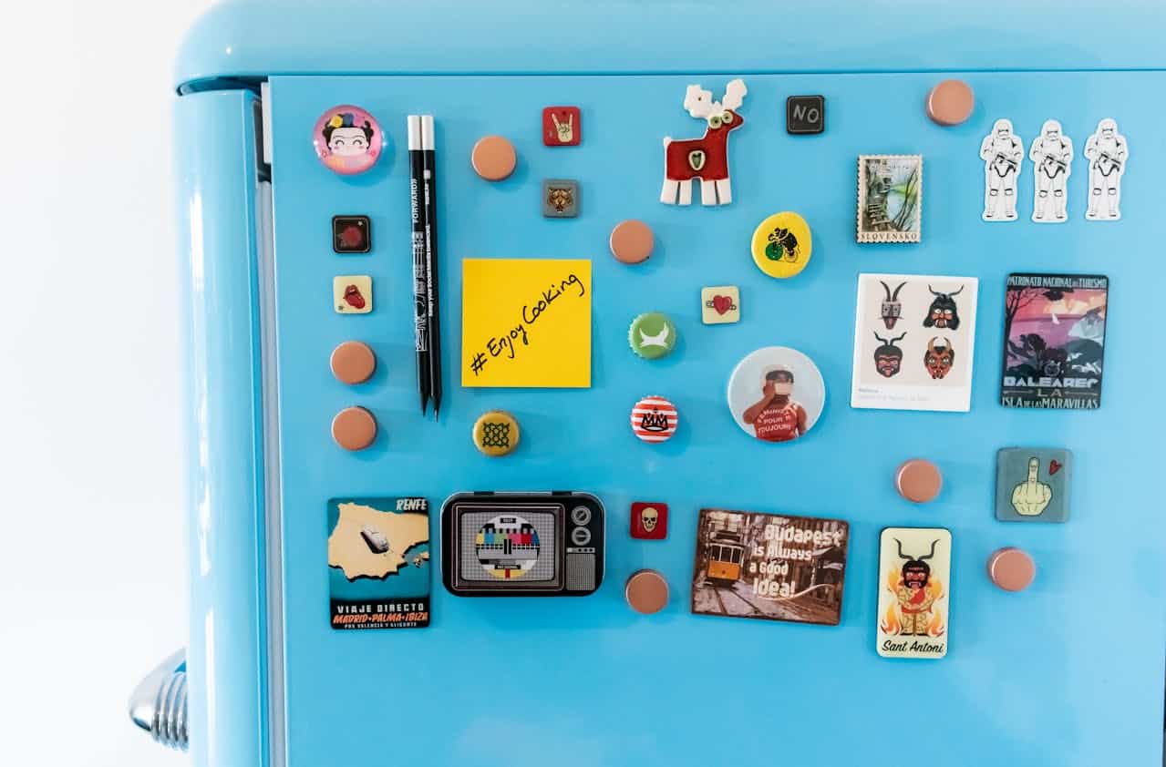 Bright blue fridge covered in colorful magnets, travel souvenirs, cartoon stickers, bottle caps, and a yellow sticky note reading "#EnjoyCooking''