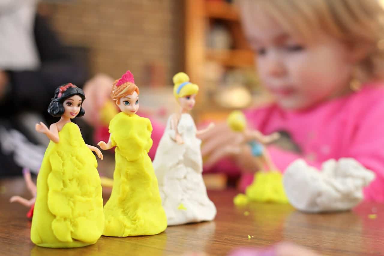 Child playing with princess dolls dressed in handmade playdough gowns, colorful modeling clay and figures on a wooden table