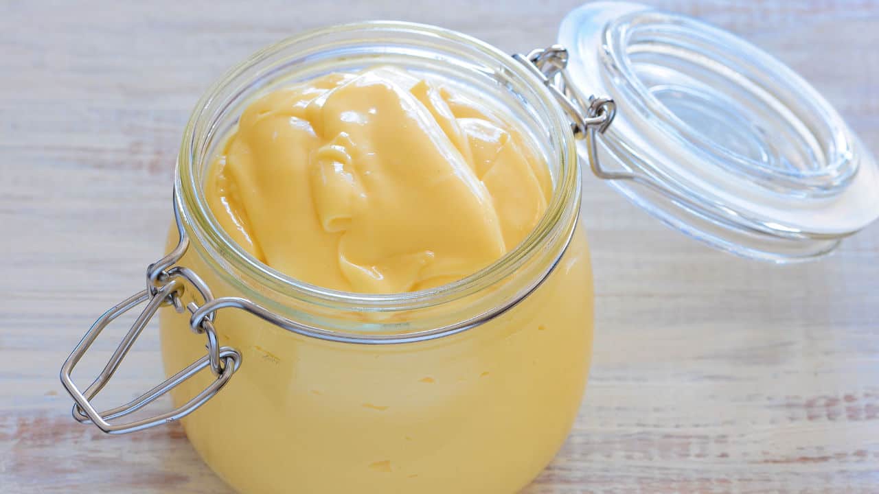 Glass jar filled with smooth, creamy yellow mayonnaise, lid open and resting on a light wooden surface