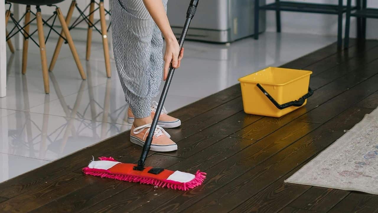 Person in gray pants using red flat mop to clean dark floor in modern interior with yellow furniture