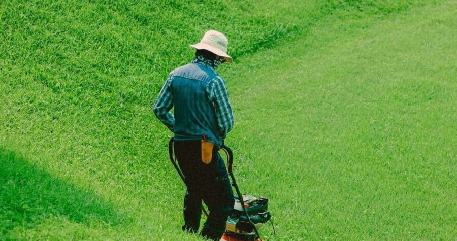 person mowing grass with a push lawn mower, wearing a hat and long-sleeved shirt, lush green lawn, slightly damp appearance, overcast lighting