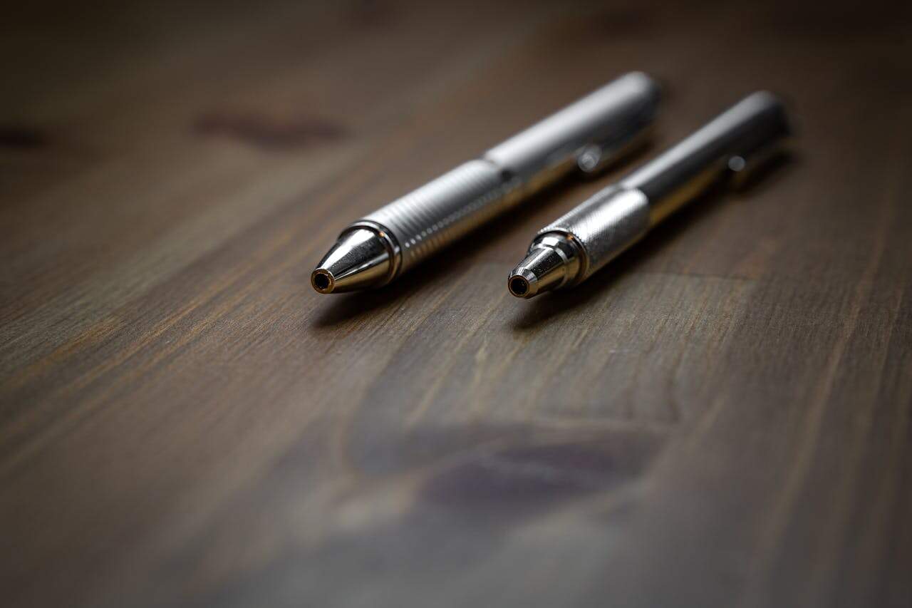 Two metallic ballpoint pens with textured grips lying on a wooden surface, close-up with shallow depth of field