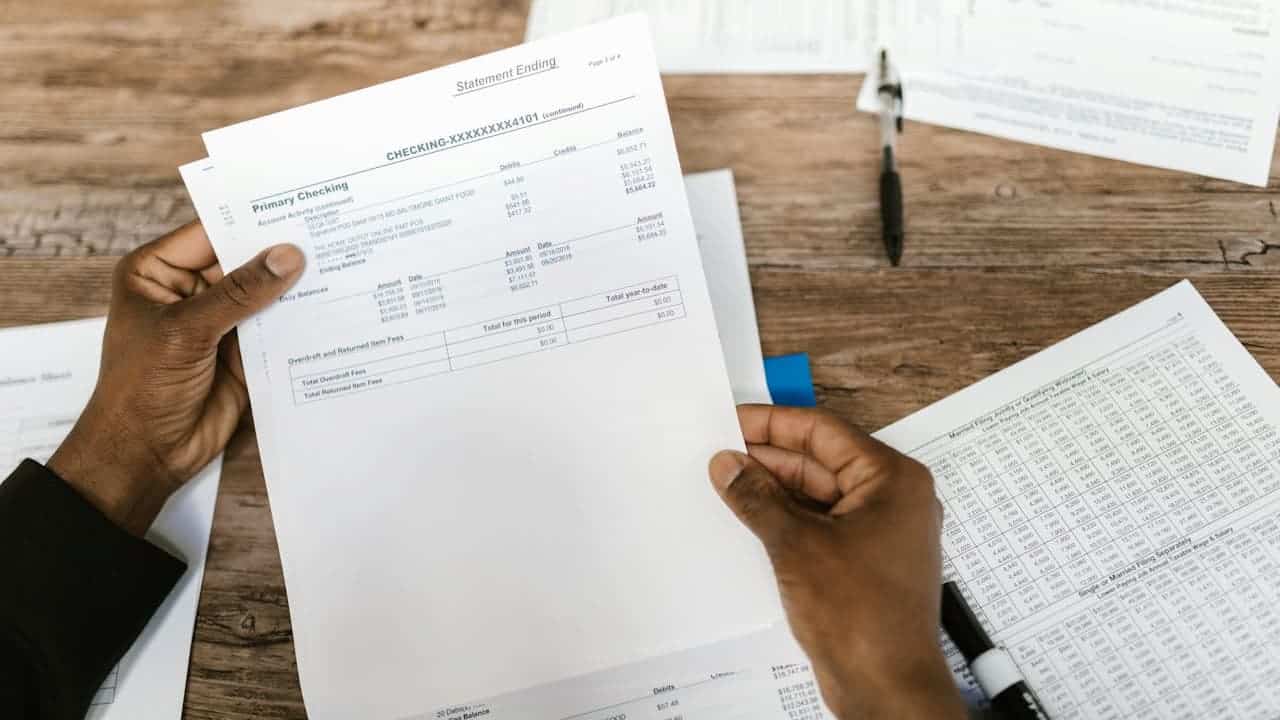 Hands holding bank statement showing checking account details, financial documents scattered on wooden desk surface, pen nearby