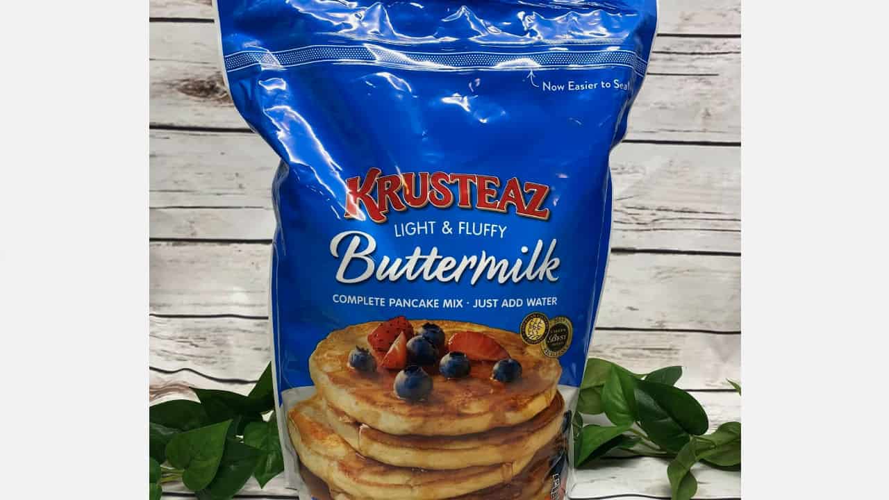 Blue Krusteaz Buttermilk pancake mix bag with a stack of pancakes topped with blueberries and strawberries on the front, placed on a wooden surface with green leaves nearby
