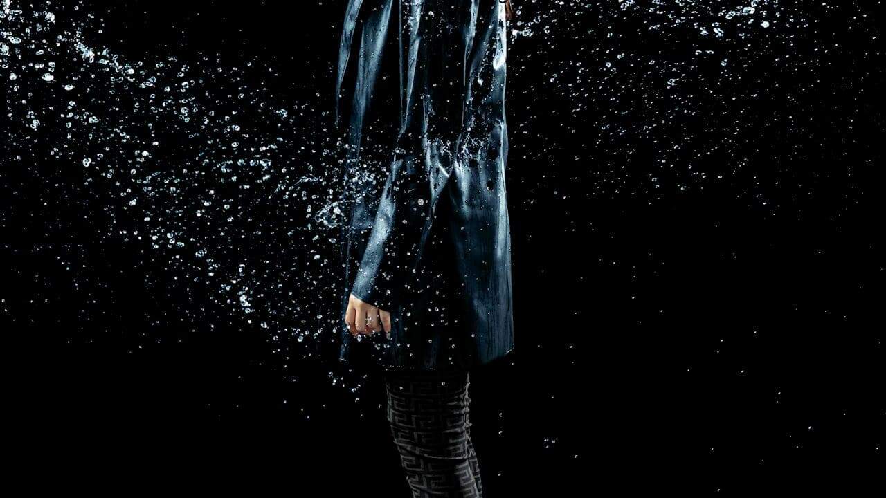 Person wearing a waterproof coat while water splashes around them, droplets in mid-air, outdoor setting, protective rain gear