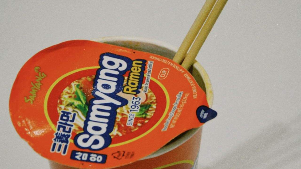 Cup of Samyang ramen with orange lid, wooden chopsticks inserted, showing noodles and broth against gray background