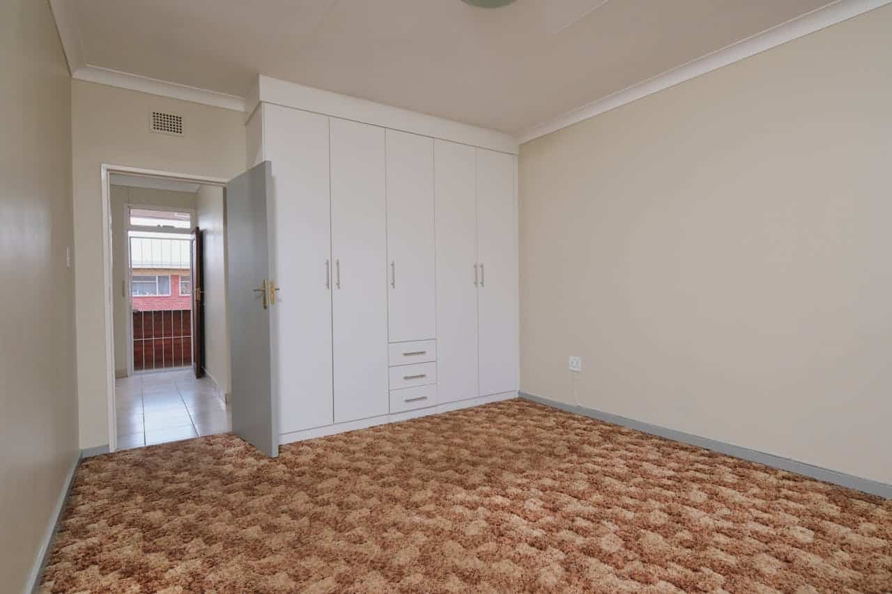 Bedroom with built-in white wardrobe, beige walls, brown patterned carpet, open door leading to hallway with tiled floor