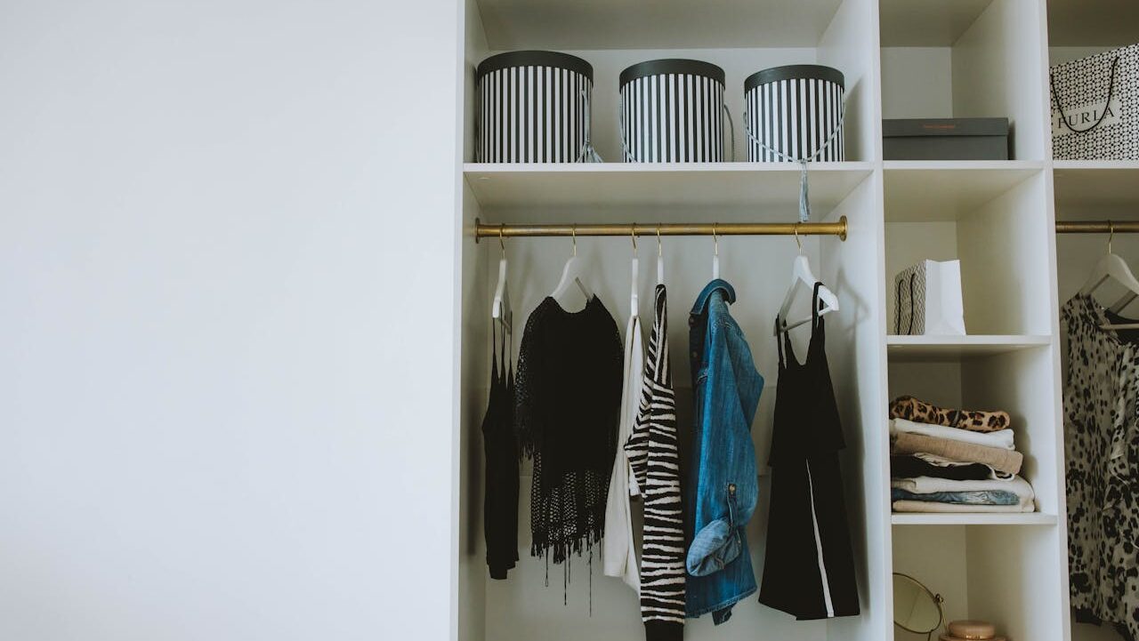 White built-in closet with hanging clothes, striped storage boxes, folded garments, and a blue denim jacket on gold rod