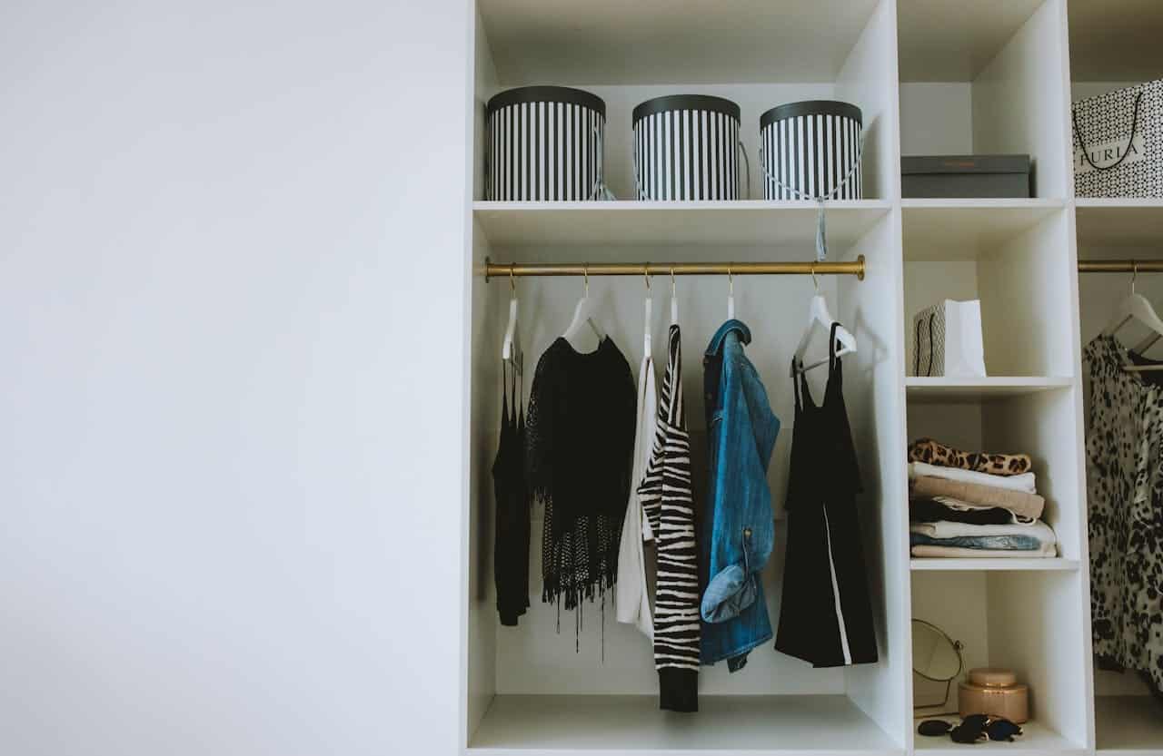 White built-in closet with hanging clothes, striped storage boxes, folded garments, and a blue denim jacket on gold rod