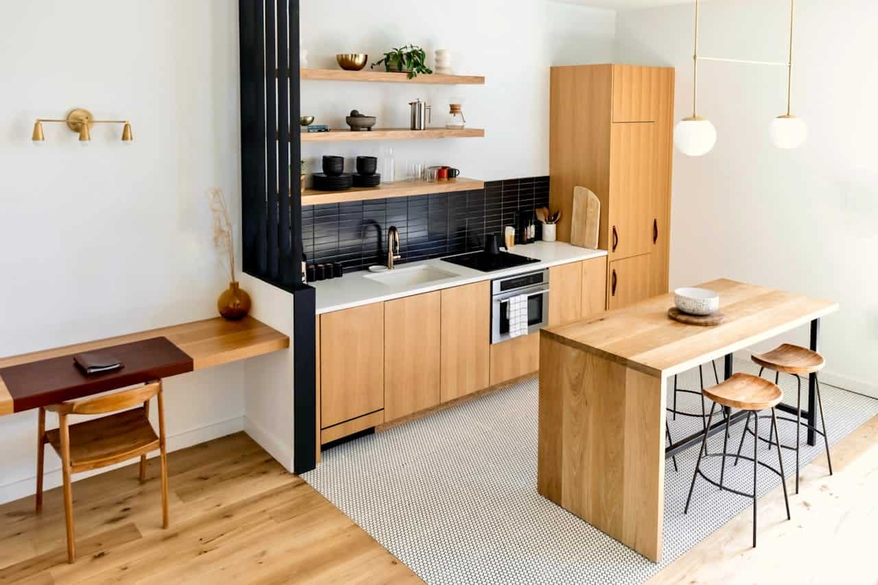 Modern Scandinavian kitchen with wooden cabinetry, black backsplash, open shelving, small desk area, wooden island with bar stools, and pendant lights