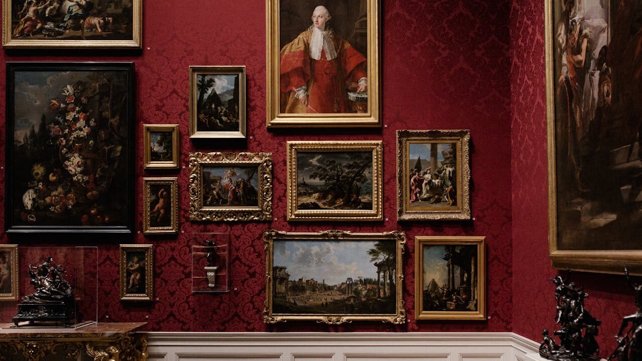 A gallery wall with various framed paintings in gold frames, featuring landscapes, portraits, and classical scenes, displayed against a rich red patterned wall