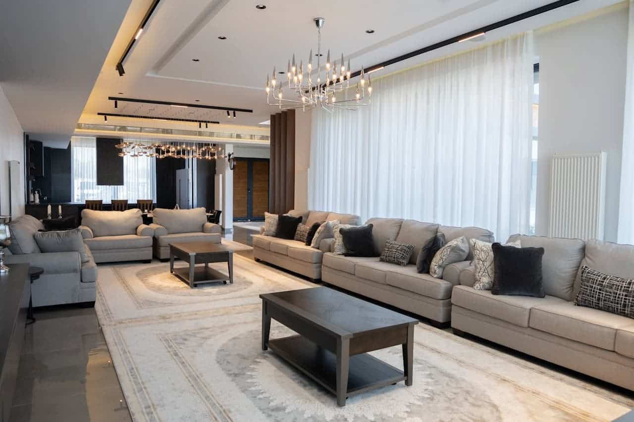 Luxurious open-concept living room with beige sofas, modern chandeliers, floor-to-ceiling white curtains, and elegant recessed ceiling design