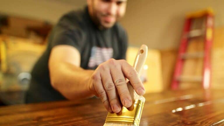 A man wearing a dark t-shirt is applying a wood stain or varnish using a wide-bristled brush, close-up focus on his hand gripping the brush