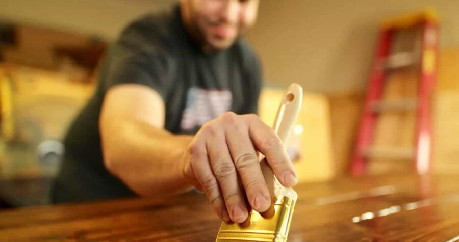A man wearing a dark t-shirt is applying a wood stain or varnish using a wide-bristled brush, close-up focus on his hand gripping the brush