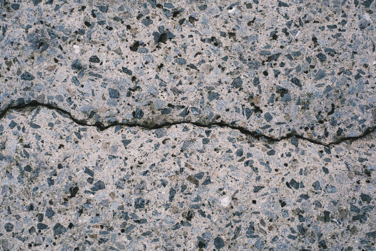 Close-up of cracked concrete surface, grey and black stone fragments embedded, uneven break running horizontally, rough texture, small air holes throughout