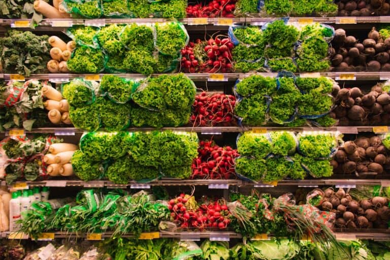 Grocery store produce section, shelves stacked with fresh leafy lettuce, red radishes, white daikon radishes, beets, and green herbs, all neatly organized and packaged in clear plastic bags, under bright lighting for display
