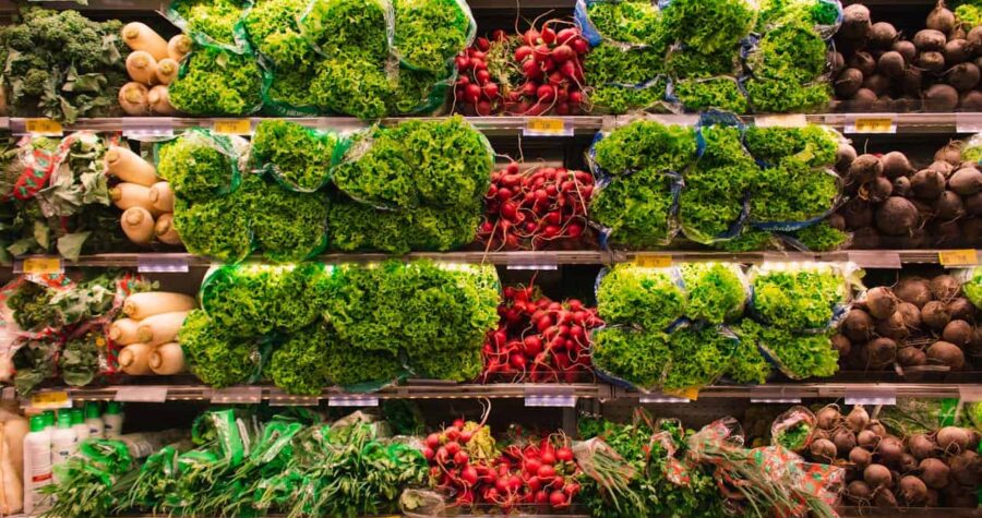 Grocery store produce section, shelves stacked with fresh leafy lettuce, red radishes, white daikon radishes, beets, and green herbs, all neatly organized and packaged in clear plastic bags, under bright lighting for display