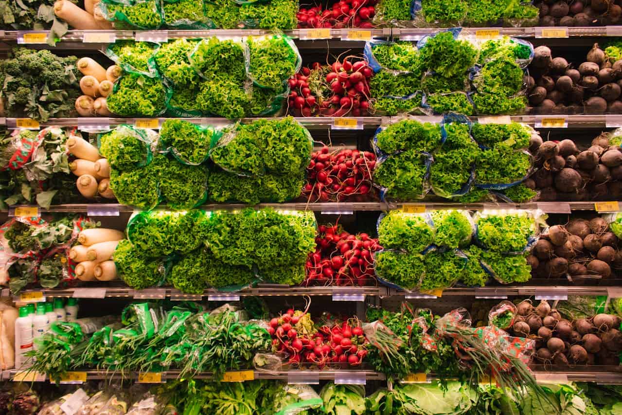 Grocery store produce section, shelves stacked with fresh leafy lettuce, red radishes, white daikon radishes, beets, and green herbs, all neatly organized and packaged in clear plastic bags, under bright lighting for display