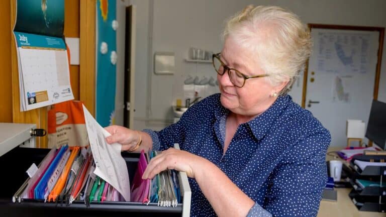 Older woman with short white hair, wearing glasses and a blue shirt, looking through a drawer filled with colorful folders and papers, office setting with a whiteboard and bulletin board in the background