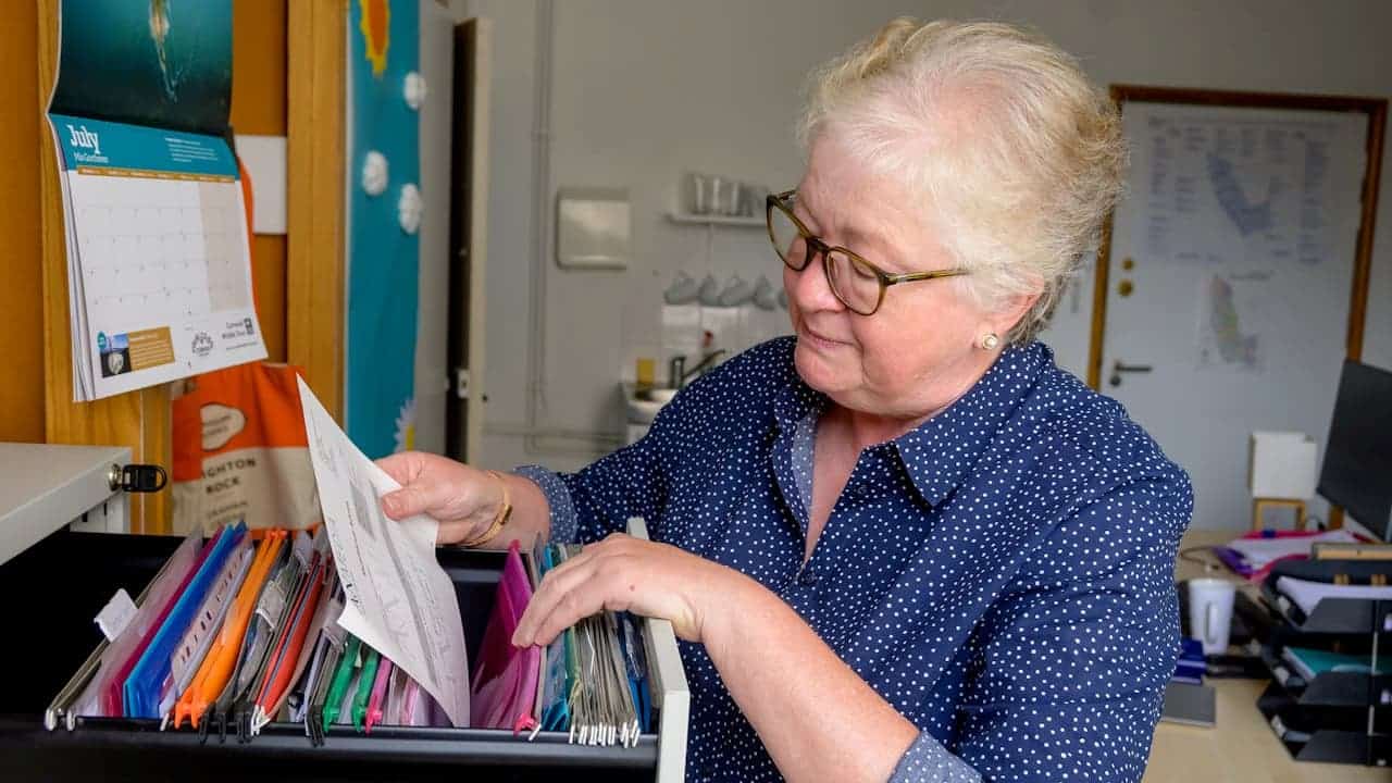 Older woman with short white hair, wearing glasses and a blue shirt, looking through a drawer filled with colorful folders and papers, office setting with a whiteboard and bulletin board in the background