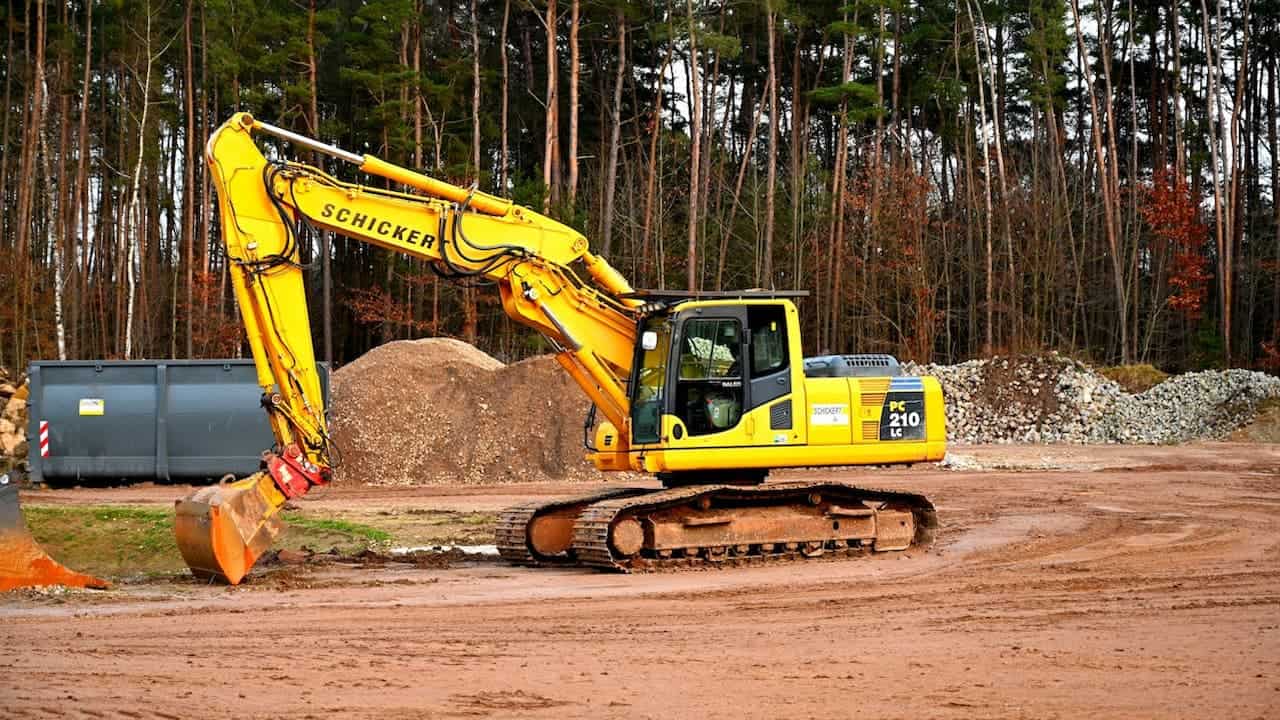Yellow Schicker excavator with bucket attachment on dirt construction site with gravel piles and pine forest background