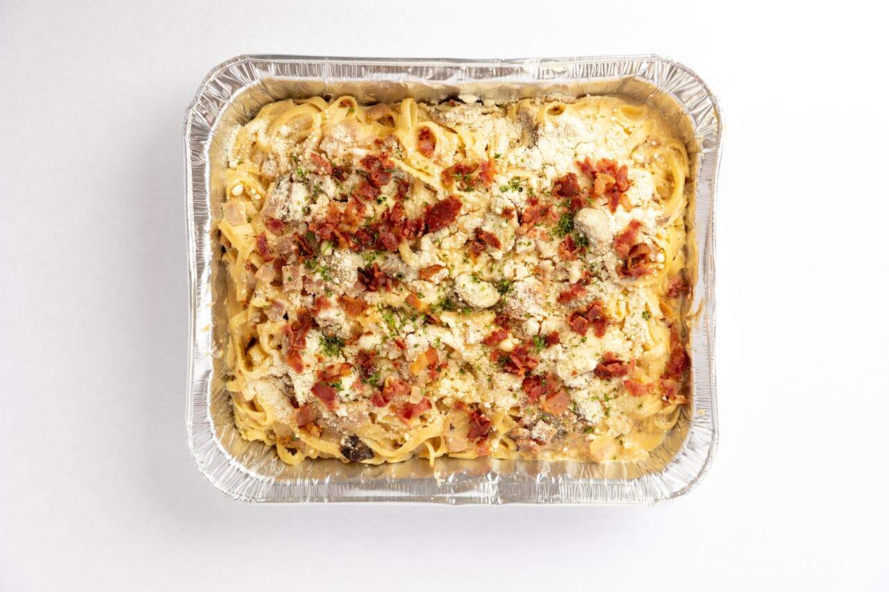 Aluminum tray filled with baked tuna noodle casserole, topped with crumbled bacon, grated cheese, and herbs, creamy sauce visible between noodles