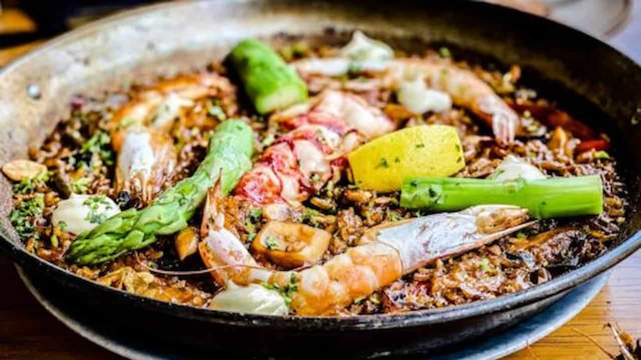 A close-up of a pan filled with seafood paella, featuring shrimp, lobster, asparagus, and lemon slices, garnished with fresh herbs, showcasing a vibrant, delicious meal with a rich blend of colors, textures, and ingredients