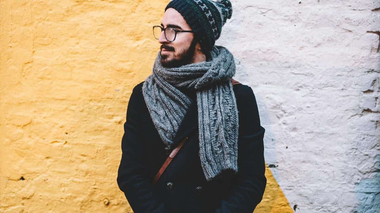 A person wearing glasses and a knitted beanie, looking to the side, draped in a silky scarf, photographed