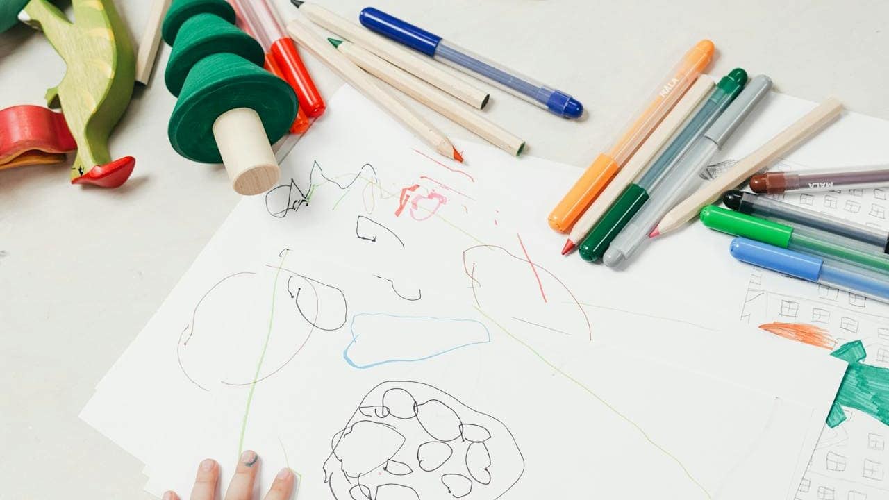 A child’s hand touches a sheet of white paper covered in colorful scribbles, surrounded by markers