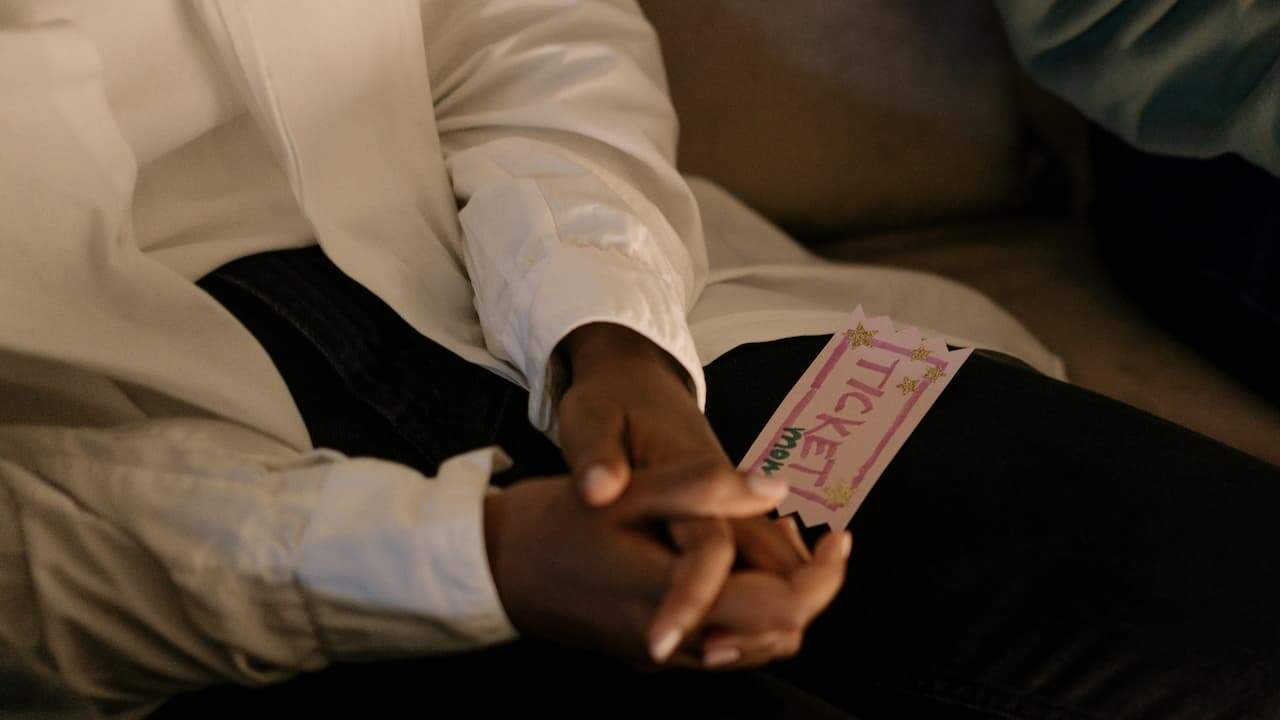Person wearing a white shirt holding a concert ticket resting on their lap, close-up view with focus on the ticket and hands, casual indoor or outdoor setting