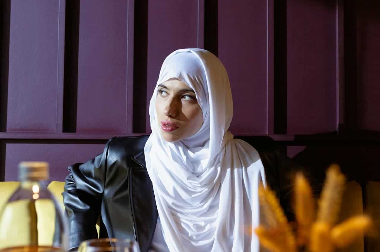 A woman in a white hijab and black leather jacket sits against a purple wall