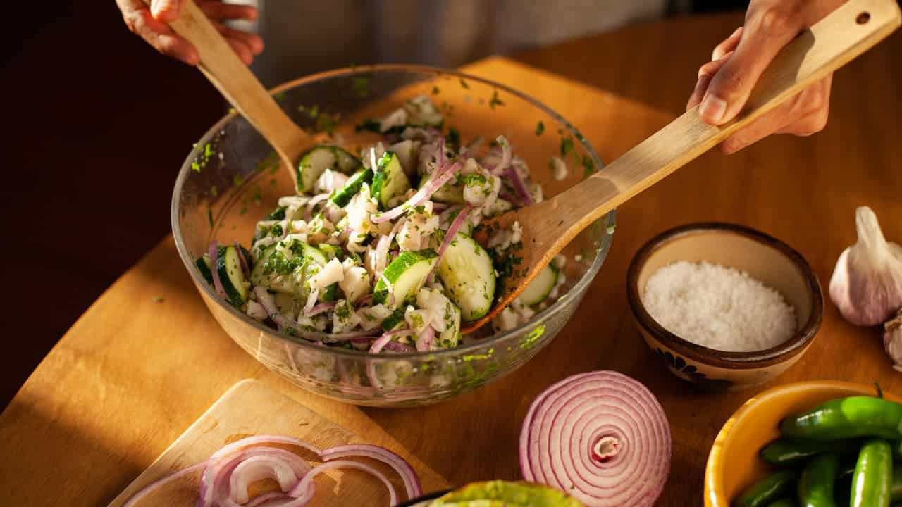 Fresh vegetable and fish salad being mixed in a clear glass tulip-shaped mixer bowl using wooden utensils, with visible ingredients like cucumber, red onion, herbs