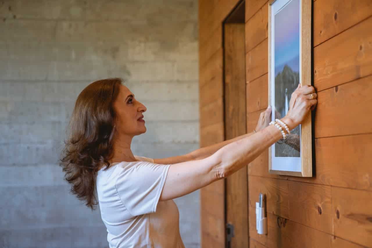 Woman in white top adjusting framed art on wooden wall, standing in profile against concrete background
