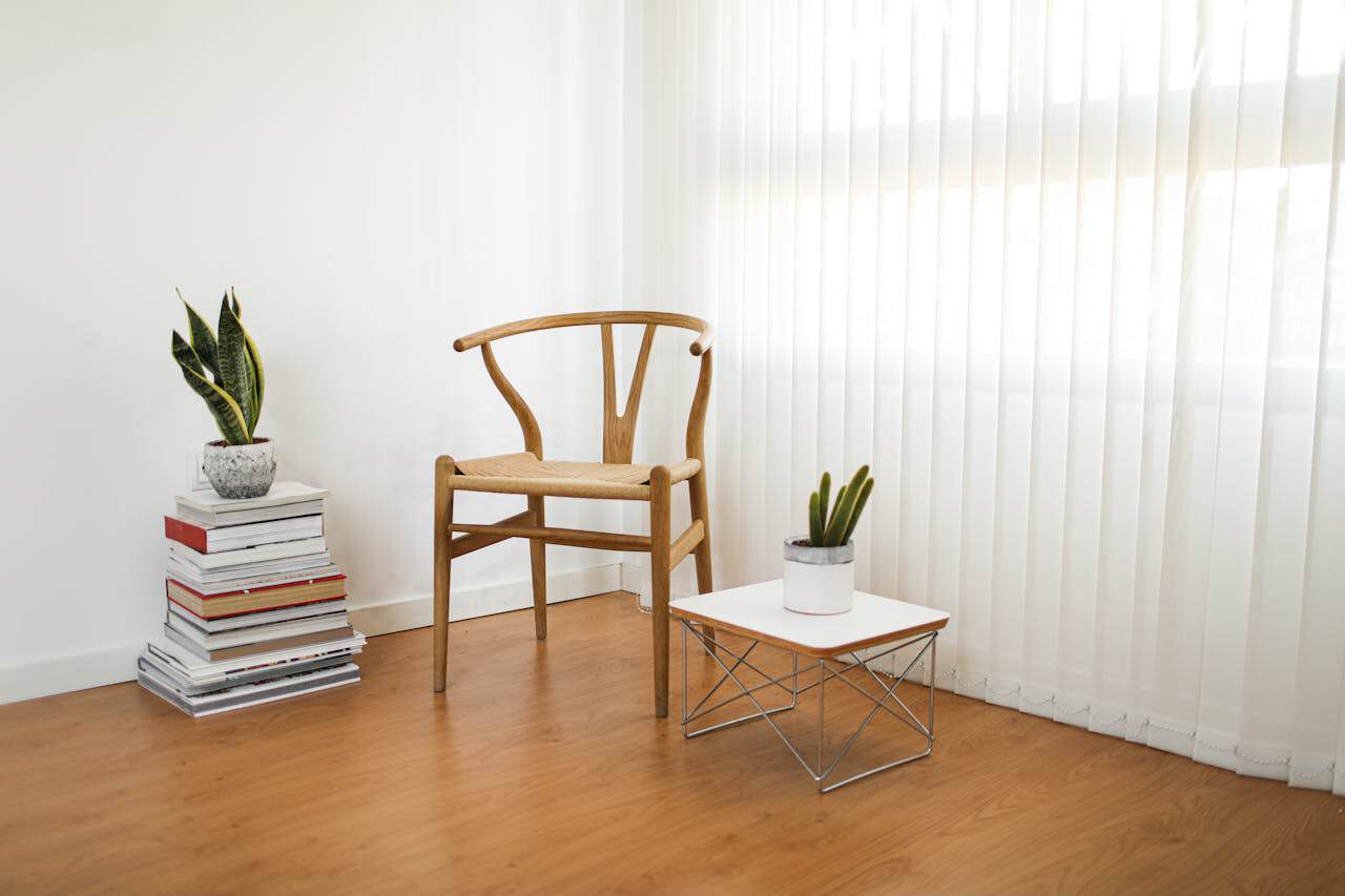 Minimalist room with wooden Wishbone chair, small side table, stacked books, two potted plants, hardwood floor, and vertical blinds