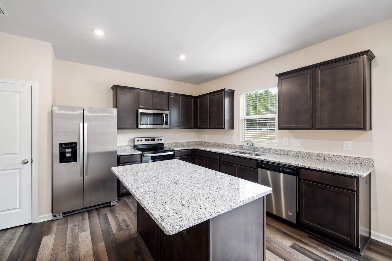 A modern kitchen with dark wood cabinetry, stainless steel appliances including a refrigerator, microwave, oven, and dishwasher, a large granite island in the center, a sink under a window with blinds, and wooden flooring, creating a clean and functional cooking space