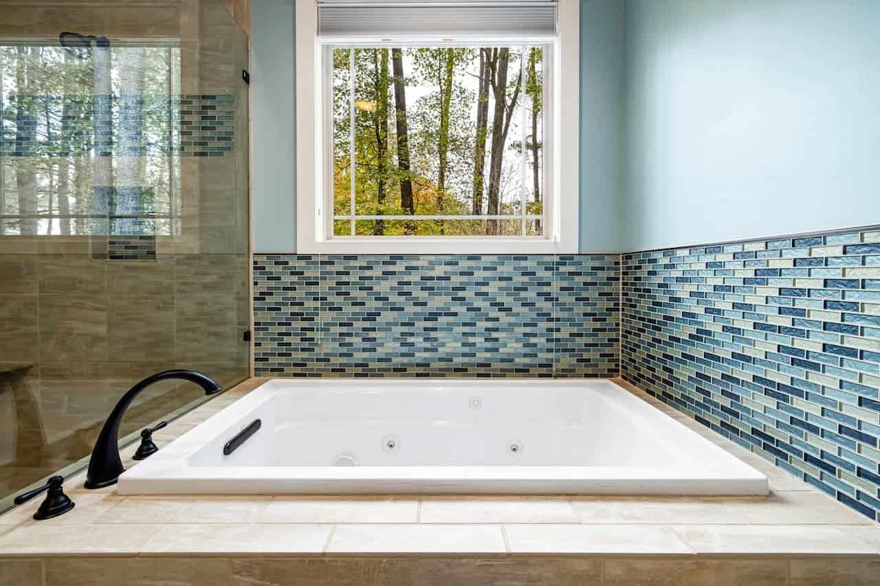 Modern bathroom with white whirlpool tub, blue glass tile backsplash, and window overlooking autumn trees
