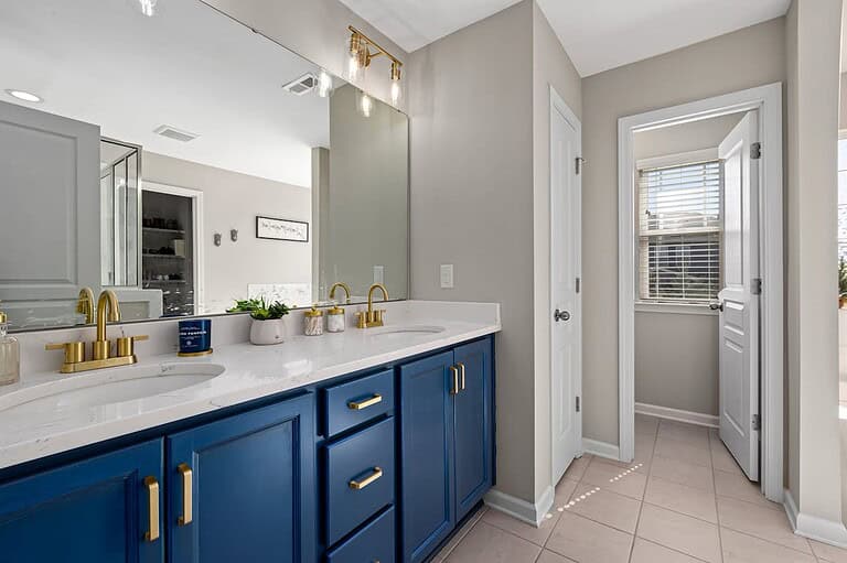 Modern bathroom with navy blue vanity cabinets, white marble countertops, gold fixtures, and beige walls leading to adjacent room