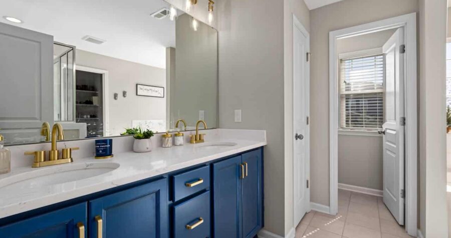 Modern bathroom with navy blue vanity cabinets, white marble countertops, gold fixtures, and beige walls leading to adjacent room