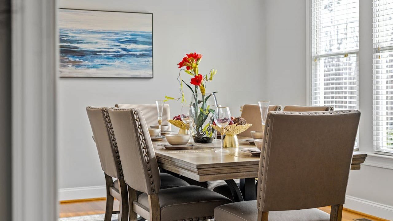 Elegant dining room with wooden table, upholstered chairs, red flower centerpiece, ocean artwork, white blinds, and ceiling light fixture