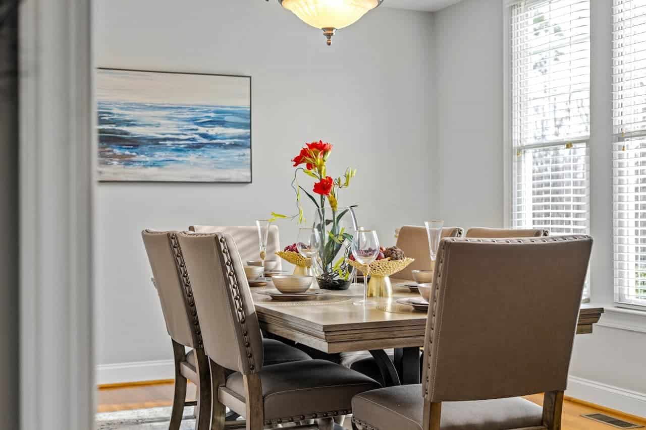 Elegant dining room with wooden table, upholstered chairs, red flower centerpiece, ocean artwork, white blinds, and ceiling light fixture