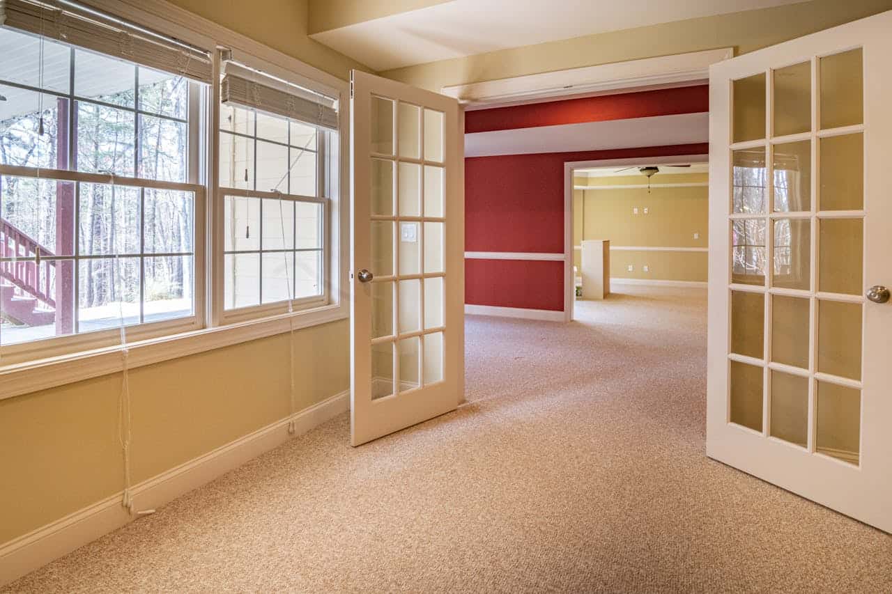 Empty room with beige wall to wall carpet, French doors, windows overlooking trees, and a red accent wall visible through doorway