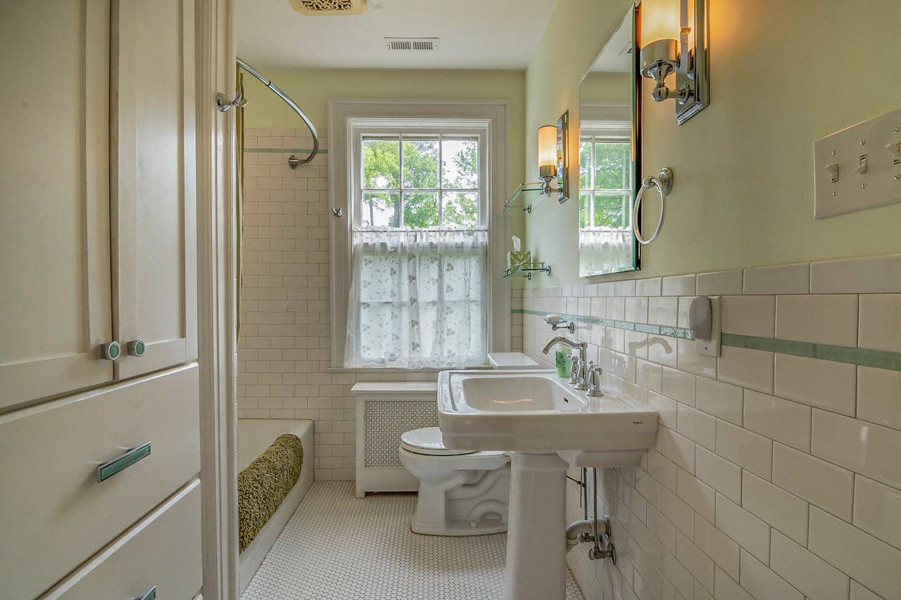 Narrow bathroom with a vintage design featuring a pedestal sink, toilet, bathtub with green towel, tiled walls and floor, window with lace curtain, and wall-mounted lighting fixtures