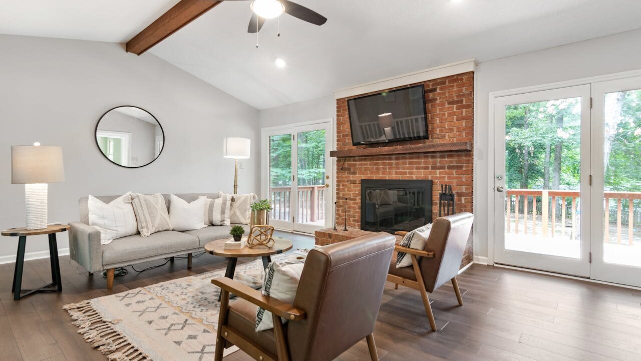Modern living room with brick fireplace, gray sofa, brown leather chairs, wooden accents, large jute area rug and glass doors overlooking wooded deck