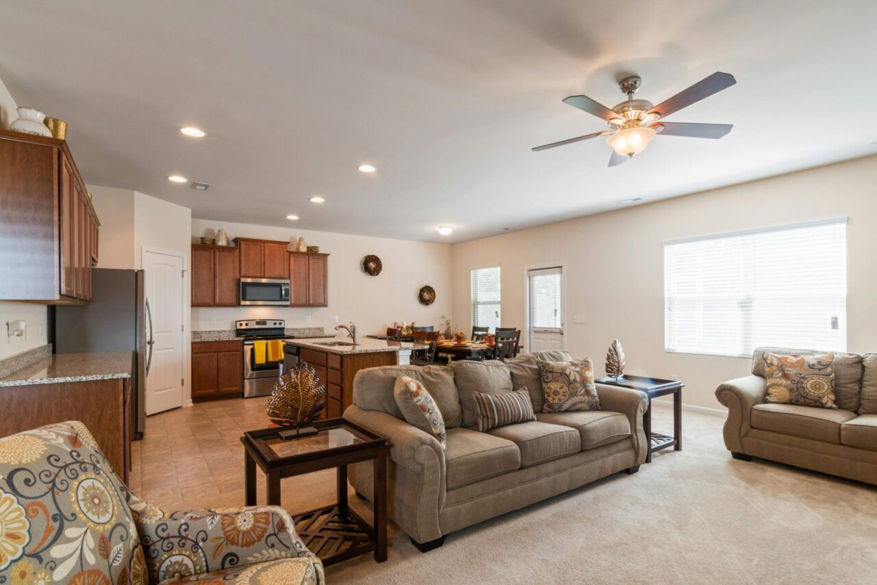 Open-concept living space, beige sofas with patterned cushions, glass-top side tables with decorative pieces, ceiling fan with lights, modern kitchen with wooden cabinets and granite countertops, stainless steel appliances, dining table in the background, large window with blinds, warm and neutral decor