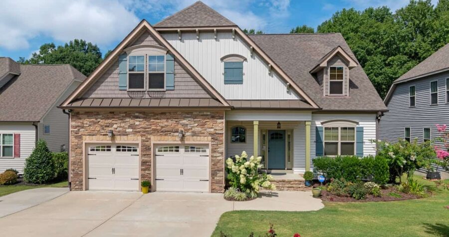 Modern suburban house with stone and siding exterior, two-car garage, landscaped yard, dormer windows, and flower-filled front garden