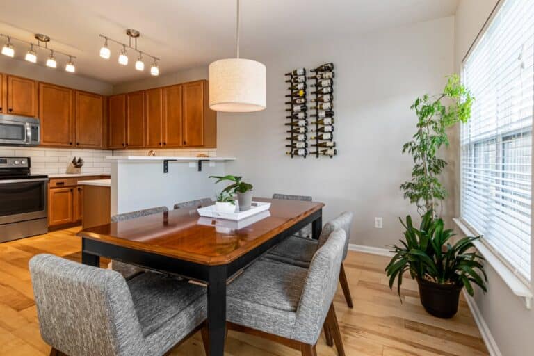 Modern kitchen-dining area with wooden table, gray chairs, white pendant light, houseplants, wine rack, and medium-toned wood cabinets