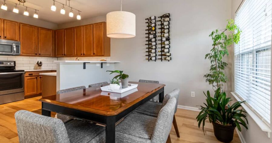 Modern kitchen-dining area with wooden table, gray chairs, white pendant light, houseplants, wine rack, and medium-toned wood cabinets