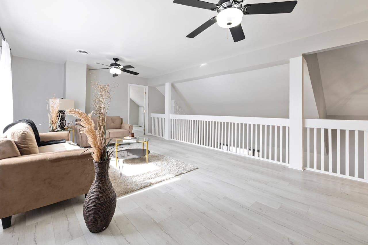 Modern loft living area with beige sofas, white railings, light wood flooring, ceiling fans, and minimalist decor