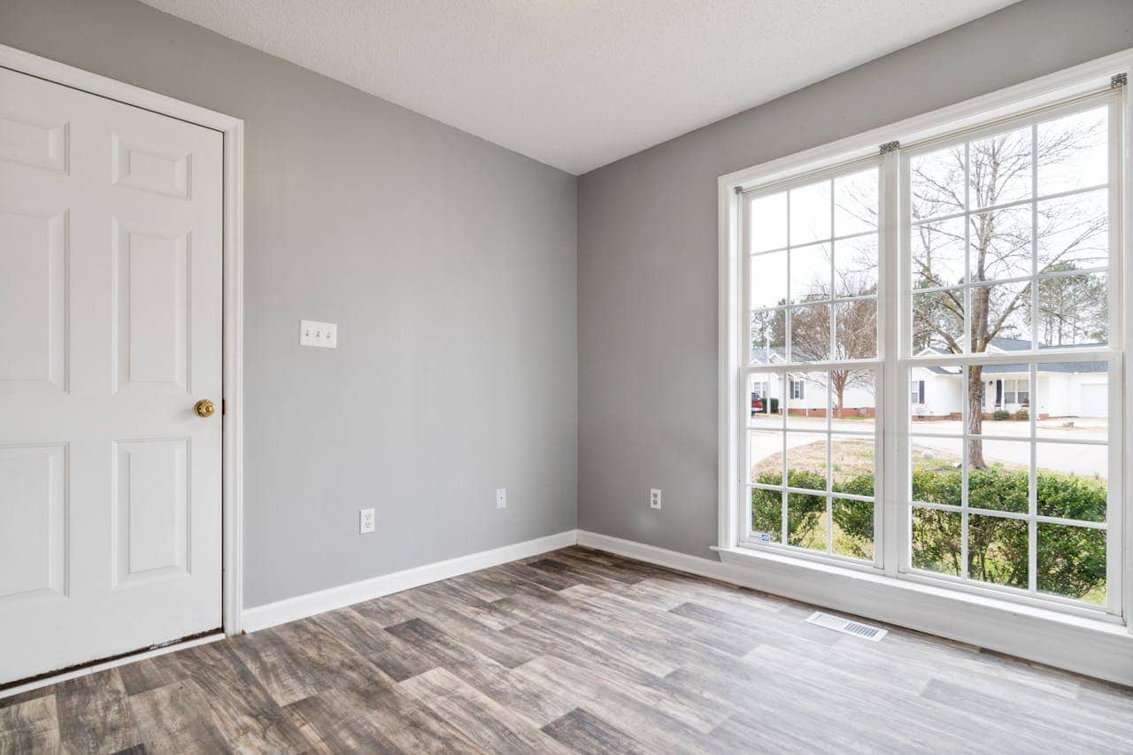Empty room with light gray walls, white trim, wood-look flooring, large window overlooking residential neighborhood, and white door