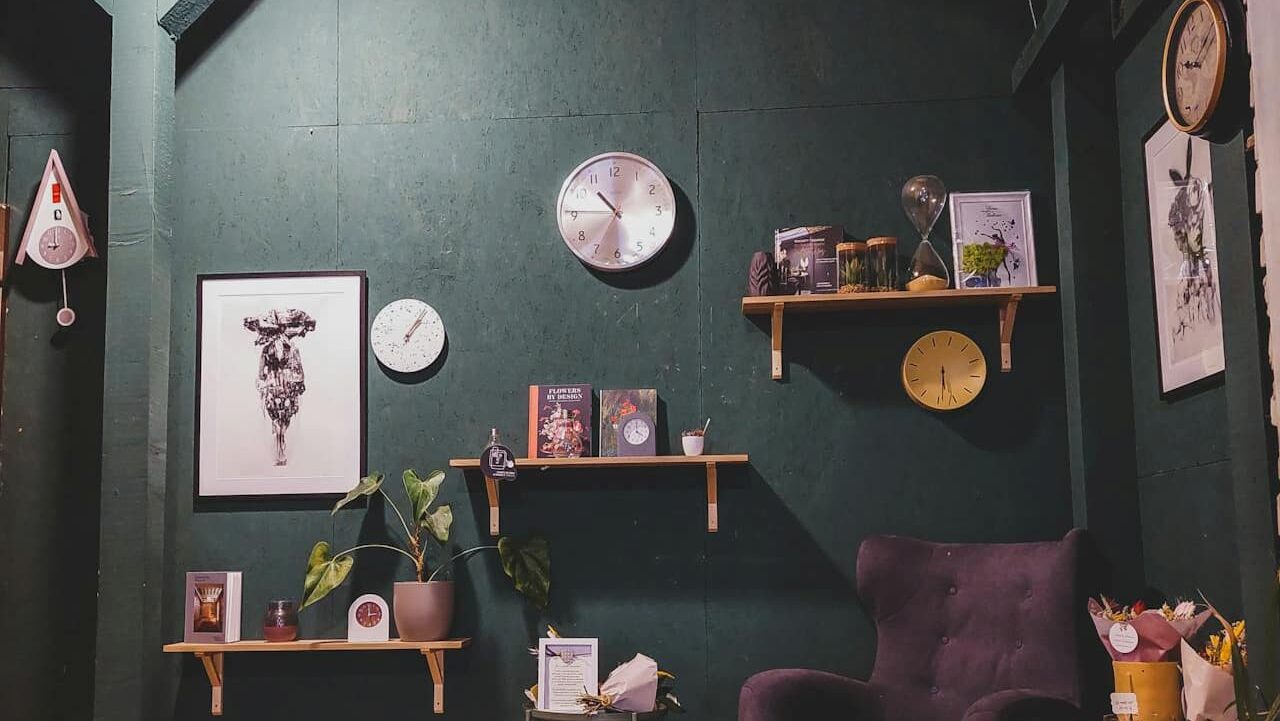 Dark green wall with multiple clocks, wooden floating shelves, framed artwork, a purple armchair, potted plants, and decorative items